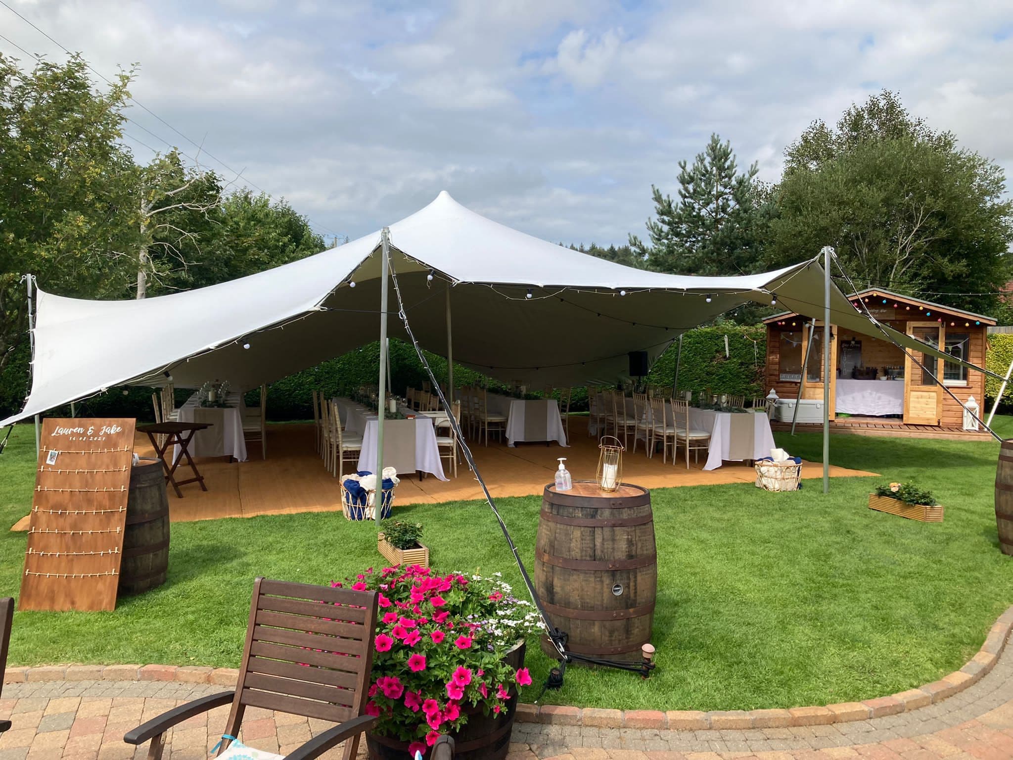 White stretch tent set up for an outdoor wedding reception on a lawn beside wooden garden buildings